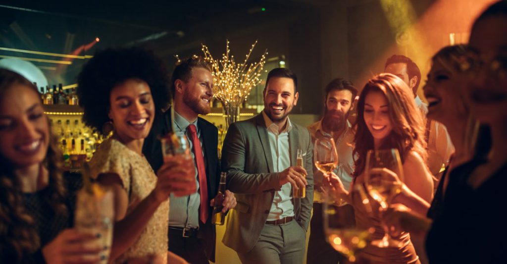 Group of five women and four men talking, laughing, and dancing at a bar event with drinks in hand.