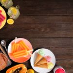 A wooden table with colorful treats, including plates of red and orange popsicles, ice cream cones, and tubs of ice cream.