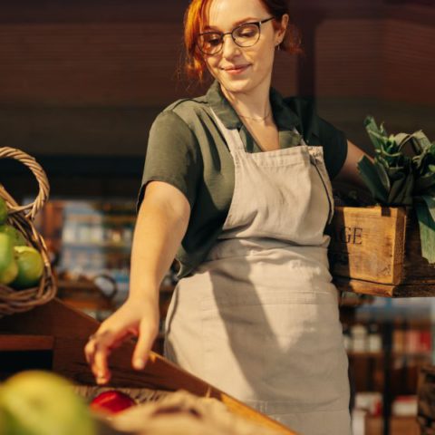 A cheery grocery store employee, wearing an apron and carrying a crate of produce, restocks the store shelves.