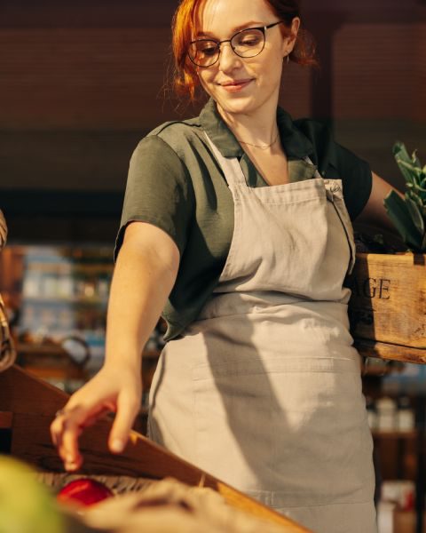 A cheery grocery store employee, wearing an apron and carrying a crate of produce, restocks the store shelves.