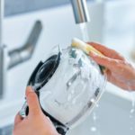 A young woman's hands holding and scrubbing a glass coffee pot carafe over her kitchen sink.
