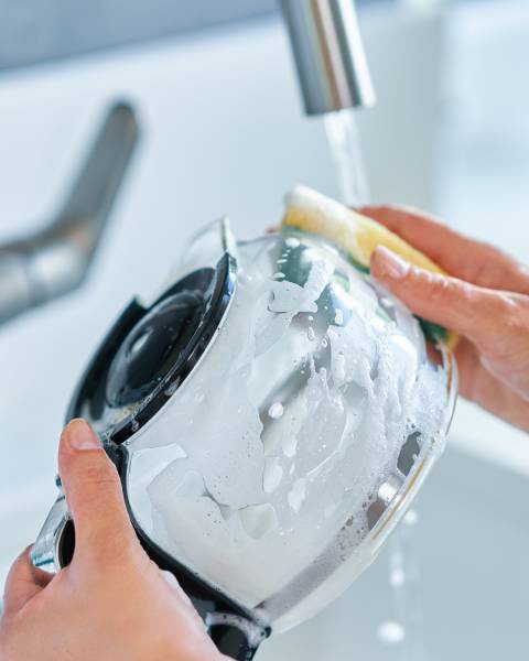 A young woman's hands holding and scrubbing a glass coffee pot carafe over her kitchen sink.