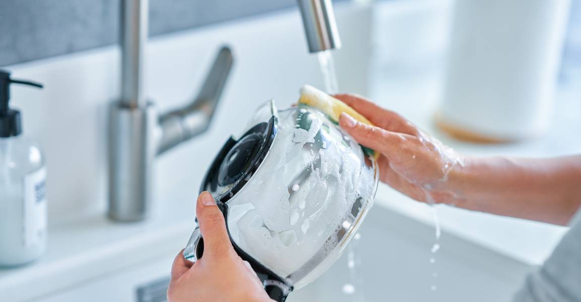 A young woman's hands holding and scrubbing a glass coffee pot carafe over her kitchen sink.