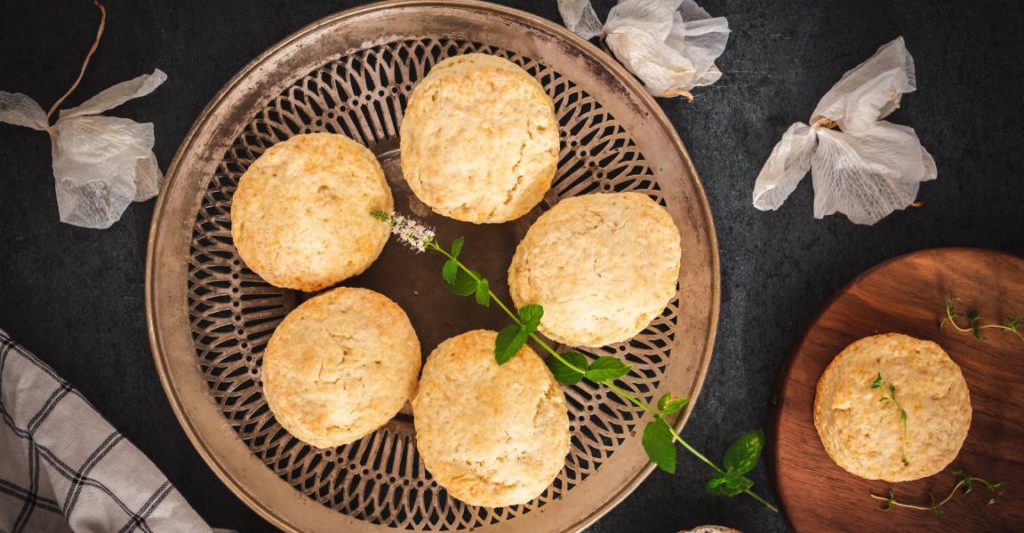 Six southern-style biscuits adorned with green garnishes are neatly arranged on a circular wooden tray.