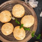 Six southern-style biscuits adorned with green garnishes are neatly arranged on a circular wooden tray.