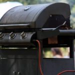 A diagonal view of a closed, outdoor grill connected to a propane tank. People are gathered in the blurred background.