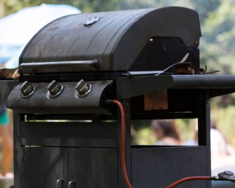 A diagonal view of a closed, outdoor grill connected to a propane tank. People are gathered in the blurred background.