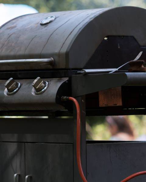 A diagonal view of a closed, outdoor grill connected to a propane tank. People are gathered in the blurred background.
