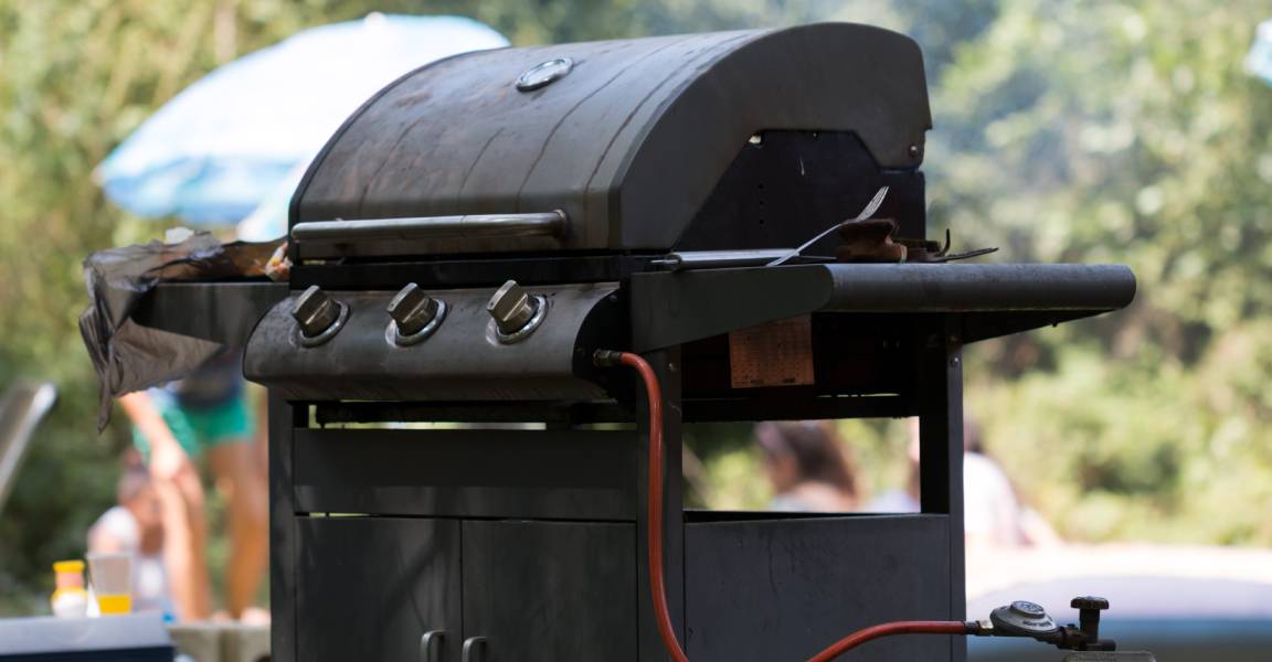 A diagonal view of a closed, outdoor grill connected to a propane tank. People are gathered in the blurred background.