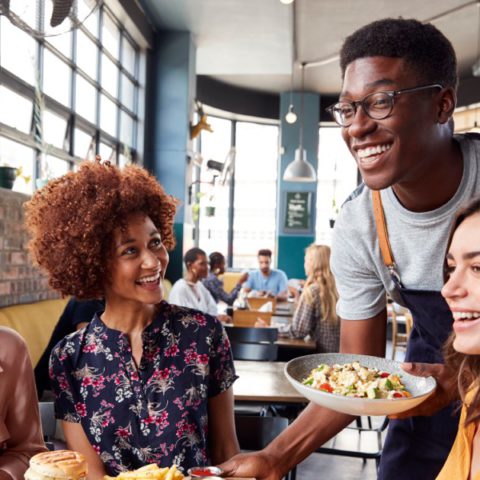 A group of young women sitting down for a meal at a restaurant. A smiling, friendly-looking waiter serves them their food.
