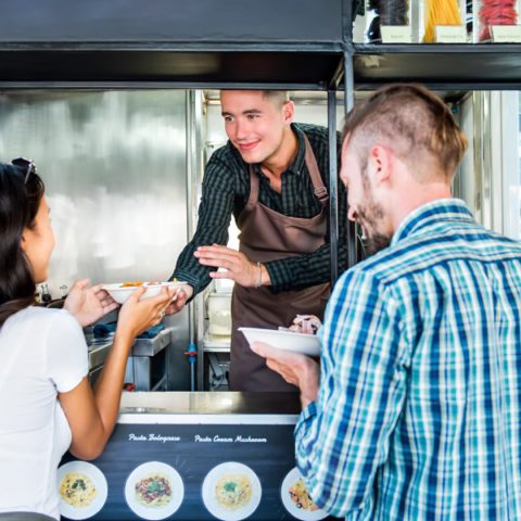 A couple picking up their orders of pasta at the window of a food truck from a worker wearing an apron.