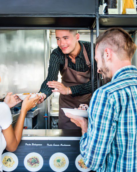 A couple picking up their orders of pasta at the window of a food truck from a worker wearing an apron.