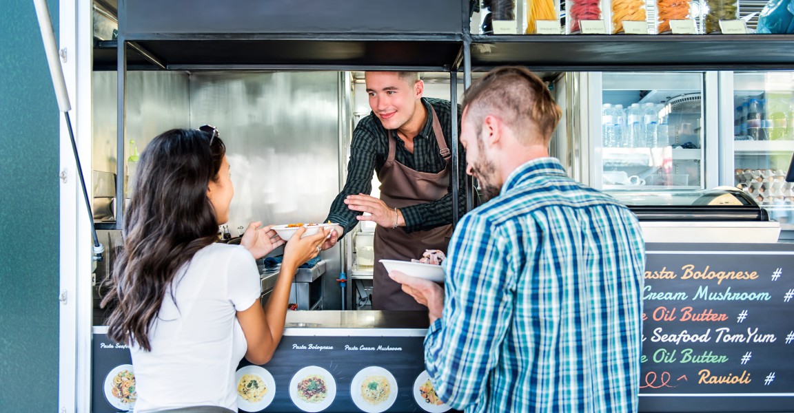 A couple picking up their orders of pasta at the window of a food truck from a worker wearing an apron.